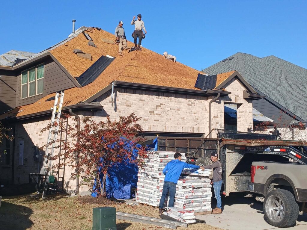 A roofing crew is actively working on a residential roof, with some sections having exposed decking and others with new underlayment. Two workers are on the roof, and two more are on the ground unloading stacks of asphalt shingles from a pallet next to a dump trailer and pickup truck.