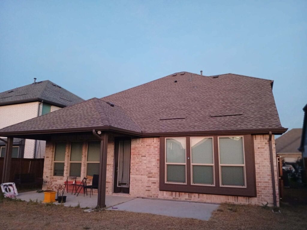 This image captures the rear view of a residential house featuring a brown asphalt shingle roof, brick exterior walls, and a covered patio area. The sky above is clear blue, suggesting either dawn or dusk.
