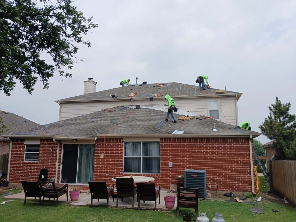 Several roofers in bright green shirts are actively working on the asphalt shingle roof of a two-story brick residential house, with some old shingles removed and new underlayment visible. The backyard patio with furniture and a grassy area is in the foreground.