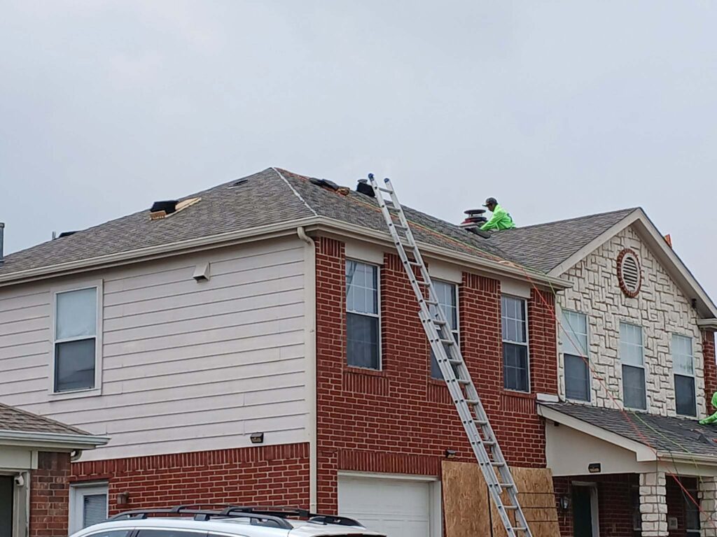 Two roofers are actively working on the asphalt shingle roof of a residential house under a cloudy sky, with a ladder leaning against the side of the house.