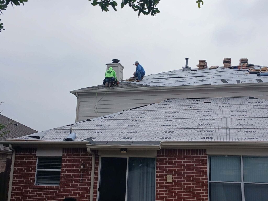 Two roofers are actively working on a residential roof, which is largely covered with synthetic underlayment. Bundles of new asphalt shingles are stacked on the roof, ready for installation.