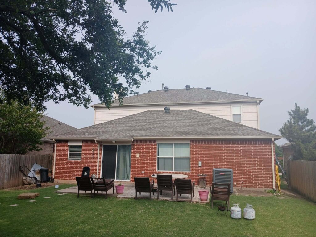 This image shows the back of a two-story residential house with a gray asphalt shingle roof, a brick first story, and light-colored siding on the second story. A patio with outdoor furniture and a grassy backyard are visible in the foreground, with a large tree partially obscuring the upper left.