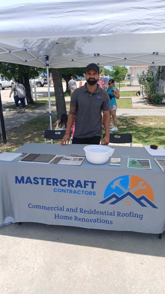 A man wearing a Mastercraft Roofing polo shirt stands behind a branded booth table displaying roofing material samples and company information at an outdoor event.
