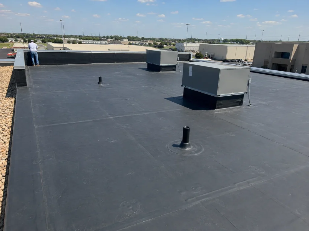 An overhead view of a large, dark gray commercial flat roof with several HVAC units and a worker standing near the edge, which is lined with gravel ballast. The background shows other commercial buildings under a clear sky.
