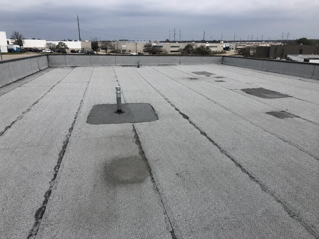 An overhead view of a commercial flat roof, showing a granulated surface with visible seams, patches, and a vent pipe. In the background, other commercial buildings and power lines are visible under a cloudy sky.