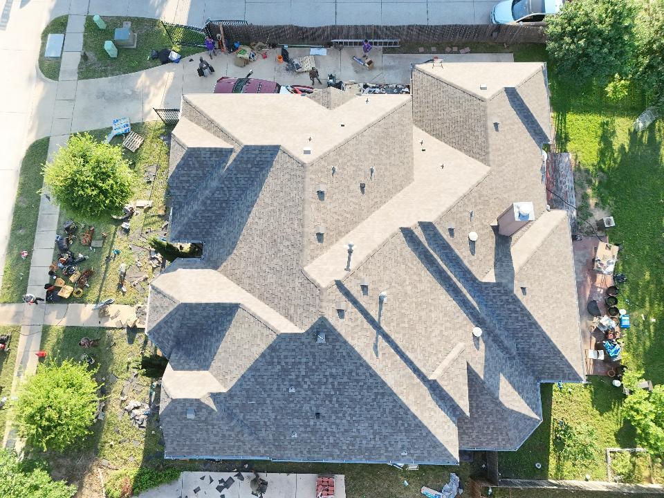 An aerial view of a residential house with a newly installed asphalt shingle roof, surrounded by debris on the ground from the roofing process. Several workers are visible on the property, managing materials and cleanup.
