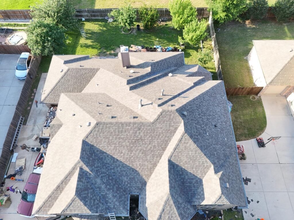 An aerial view of a residential roof, mostly covered with new asphalt shingles, showing some areas still under construction or recently completed. Construction materials and debris are visible around the house, and a worker is present on the ground.