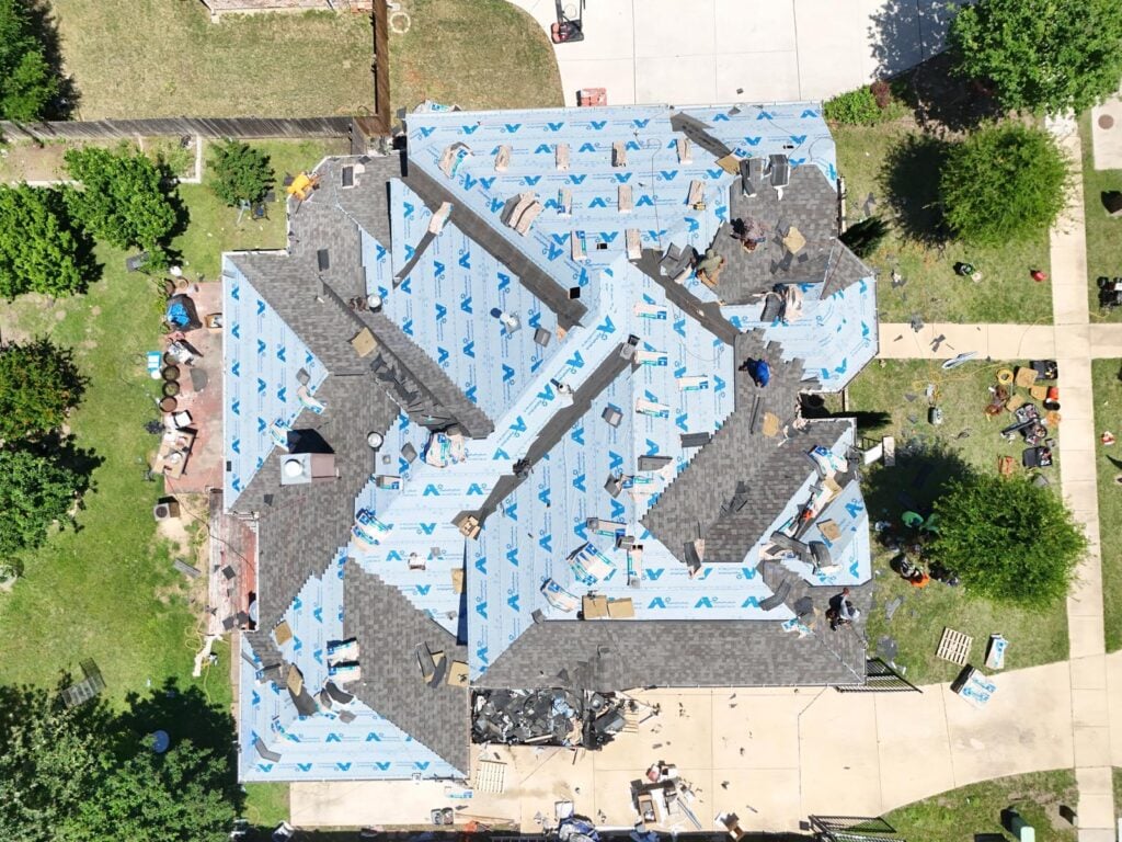 An aerial view of a residential roof undergoing replacement, with sections covered in blue underlayment and other sections already having new asphalt shingles installed. Several crew members are visible working on different parts of the roof.