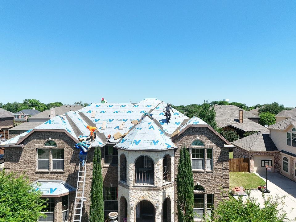 An aerial view of a large brick house with a complex roof structure, where several roofers are actively working on installing blue synthetic underlayment. One roofer is visible on a ladder, with a clear blue sky overhead.