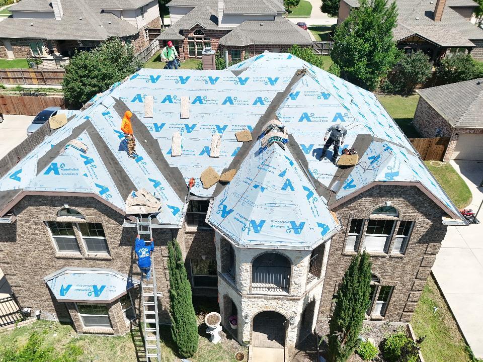 An aerial view of a large residential house with a roofing crew actively installing new underlayment and preparing for shingle installation. Several workers are visible on the roof and a ladder, with the blue underlayment covering most of the roof surface.