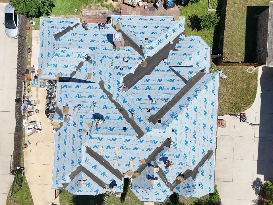 An aerial view of a residential roof during construction, showing blue synthetic underlayment installed across most of the roof, with several workers actively engaged in the process. Debris and materials are visible on the ground next to the house.