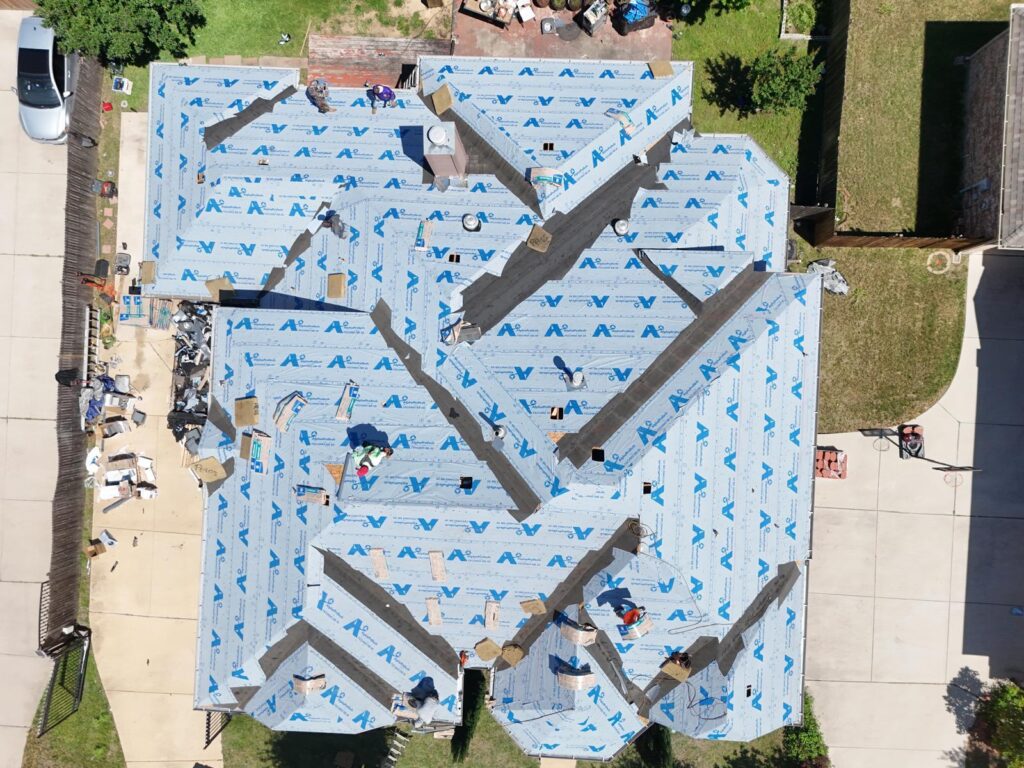 An aerial view of a residential roof under construction, with blue synthetic underlayment installed across most of the roof and several workers actively working. Debris and materials are visible on the ground around the house.