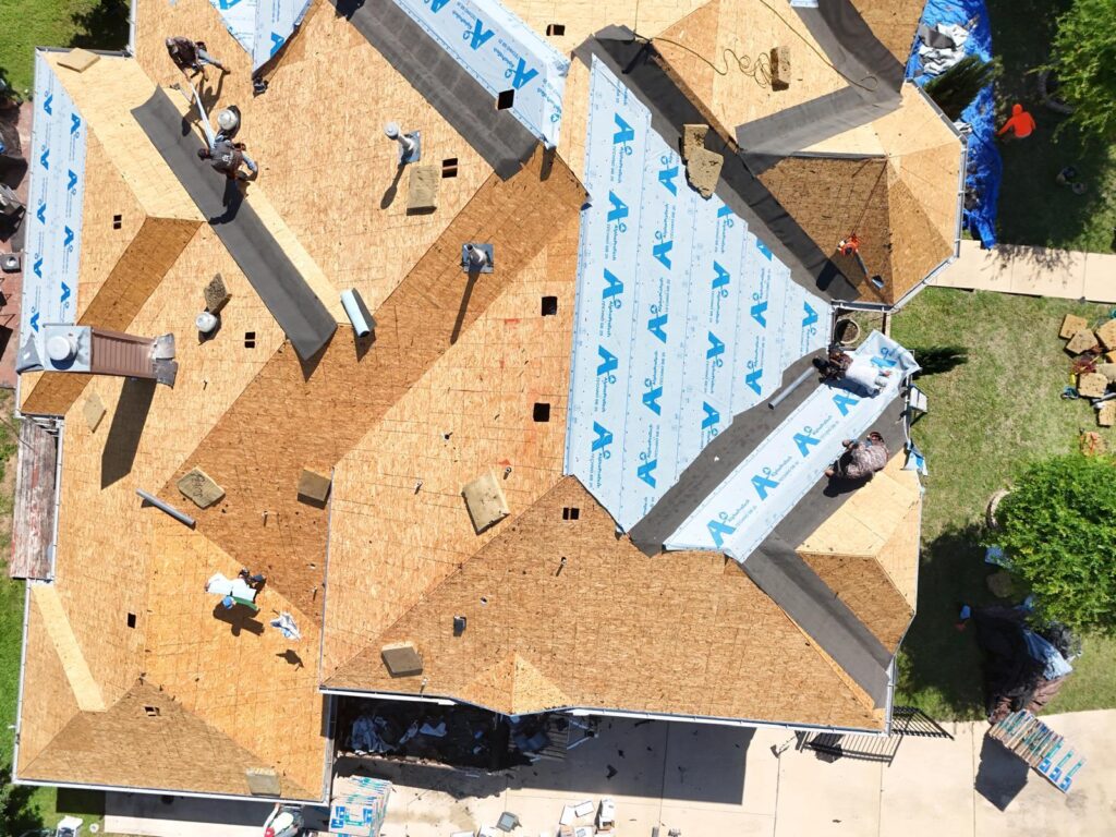 An aerial view of a residential roof undergoing replacement, with multiple crew members actively installing underlayment over exposed plywood decking. Various roofing materials and tools are scattered across the roof and surrounding ground.