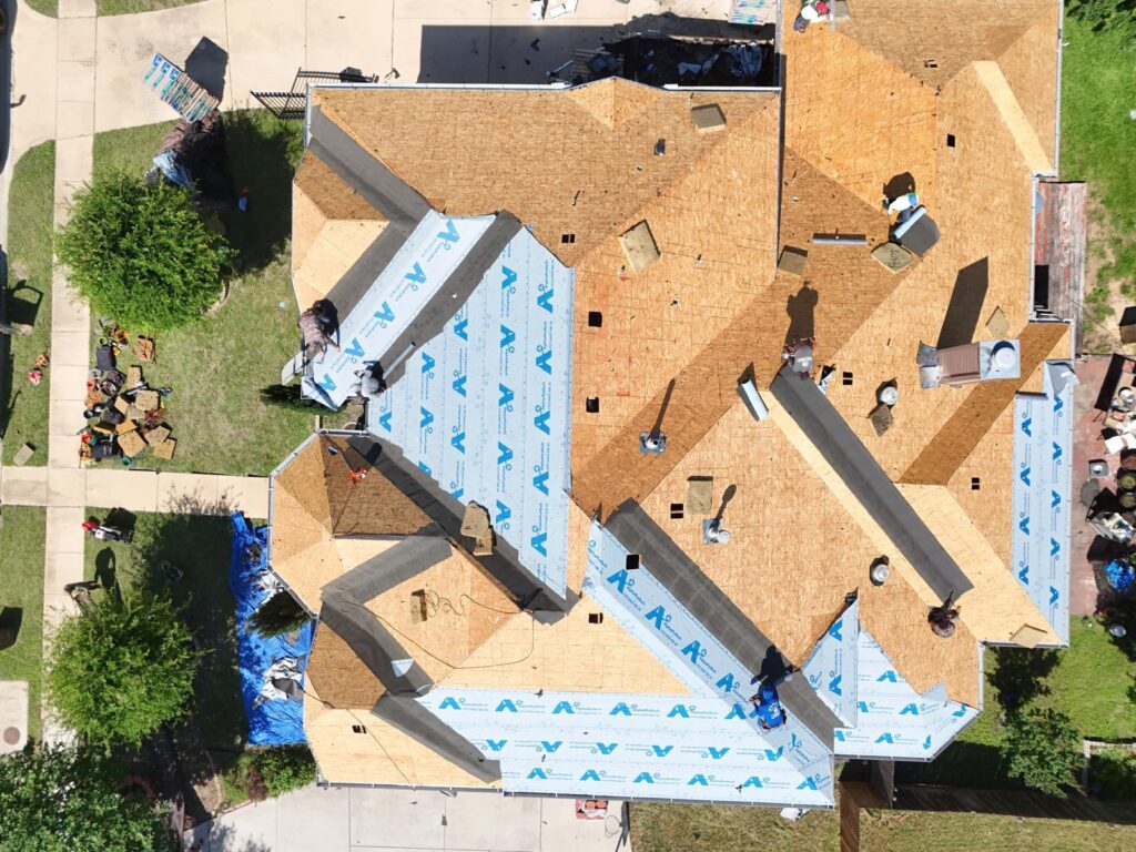 An aerial view of a residential roof undergoing replacement, showing exposed plywood decking and partially installed synthetic underlayment with several crew members actively working.