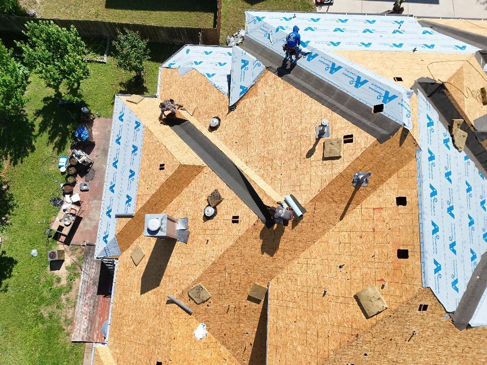 An aerial view shows a residential roof under construction, with several crew members installing blue synthetic underlayment over the plywood decking. Roofing materials and tools are scattered across the roof and on the ground next to the house.