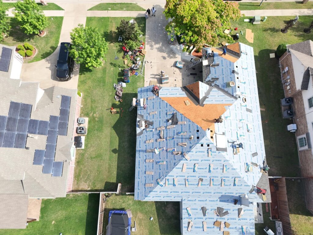 An aerial view of a residential roof undergoing replacement, with blue underlayment installed and bundles of asphalt shingles being laid. Several crew members are visible working on different sections of the roof.