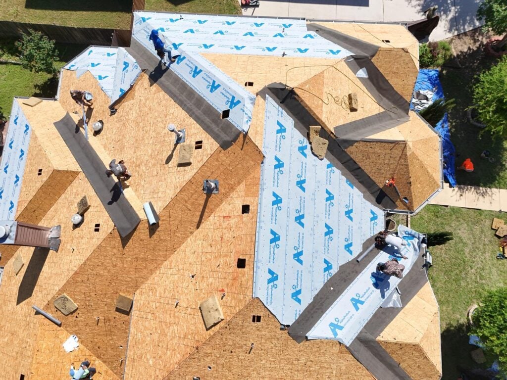 An aerial view of a residential roof under construction, with several crew members actively installing underlayment over the exposed plywood decking. Sections of blue AlphaProtech underlayment are visible, along with various tools and materials.
