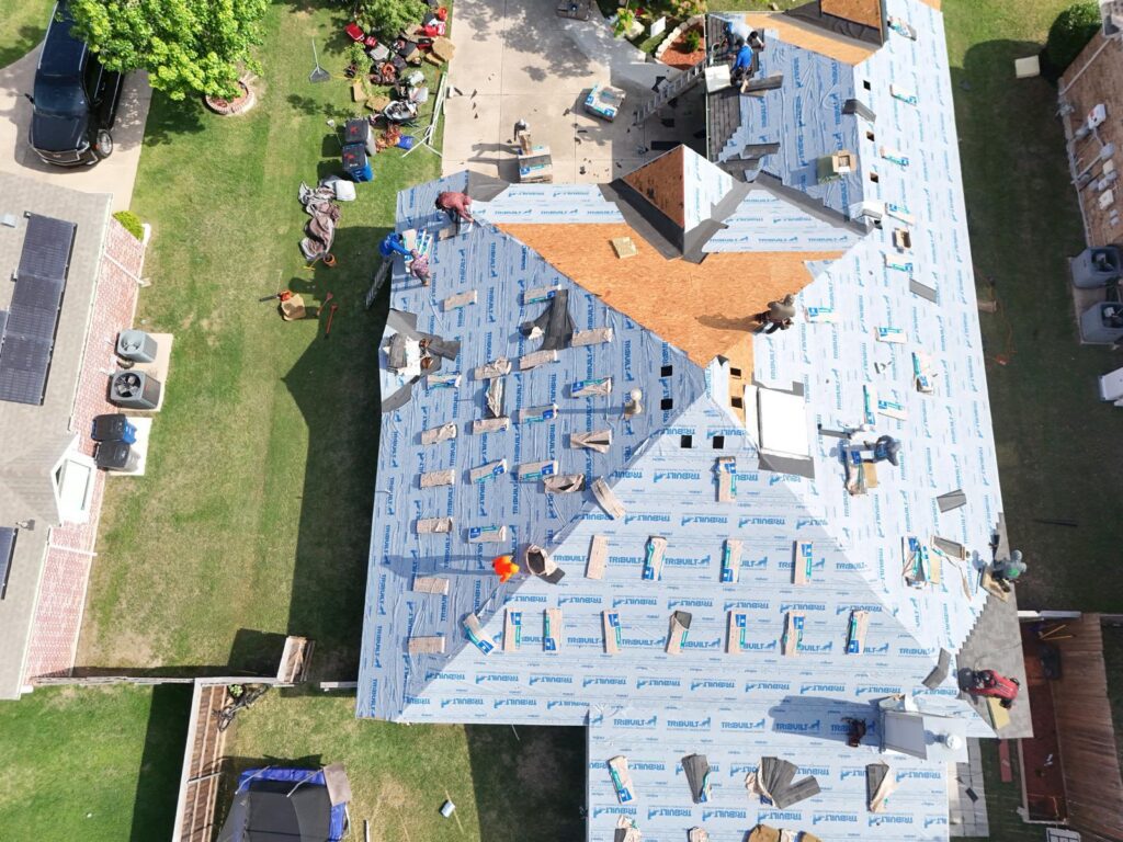 An aerial view of a residential roof undergoing replacement, with crew members installing blue synthetic underlayment and some initial bundles of asphalt shingles visible on the roof deck. The surrounding yard shows some construction debris and a parked vehicle.