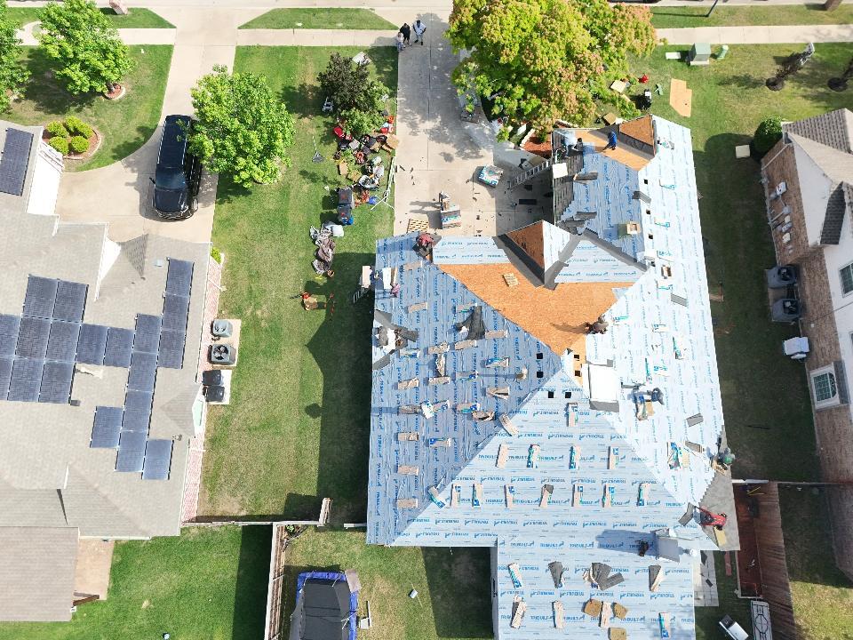 An aerial view of a residential roof undergoing replacement, with blue underlayment installed over most of the decking and several workers actively engaged in the roofing process. Debris and equipment are visible on the ground around the house.