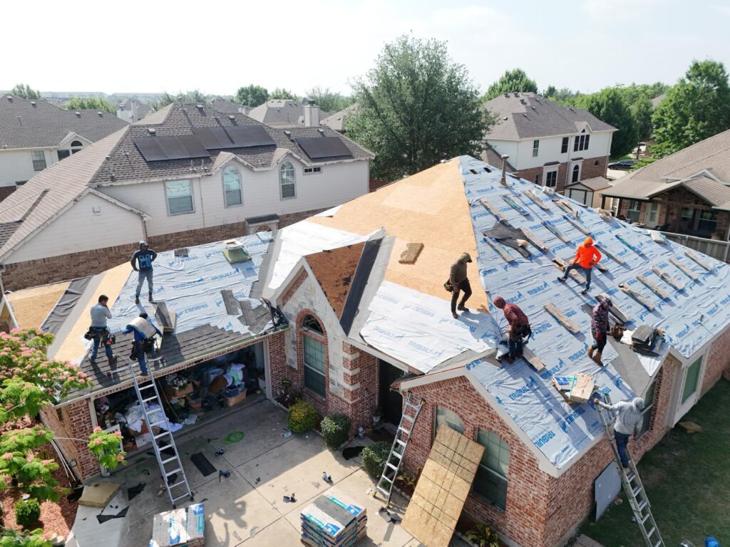 An aerial view captures a residential house undergoing roof replacement, with multiple roofers actively working on various sections of the roof, which shows exposed plywood, underlayment, and stacked new shingles.