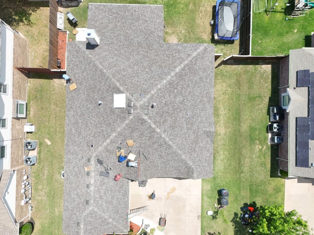 An aerial view of a residential roof undergoing replacement, with several crew members visible on the roof and ground, and roofing materials scattered around.