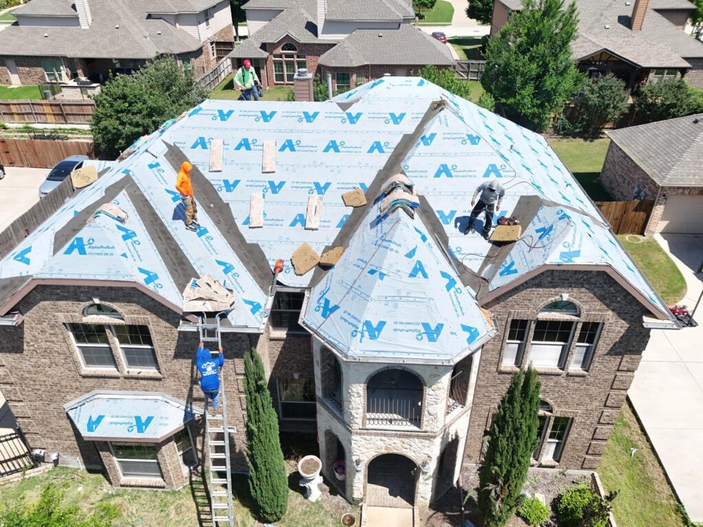 An aerial view of a large residential house with multiple roofers actively working on installing new underlayment and preparing for shingle installation. The roof is mostly covered with blue AlphaProtech underlayment, and several crew members are visible on different sections of the roof.