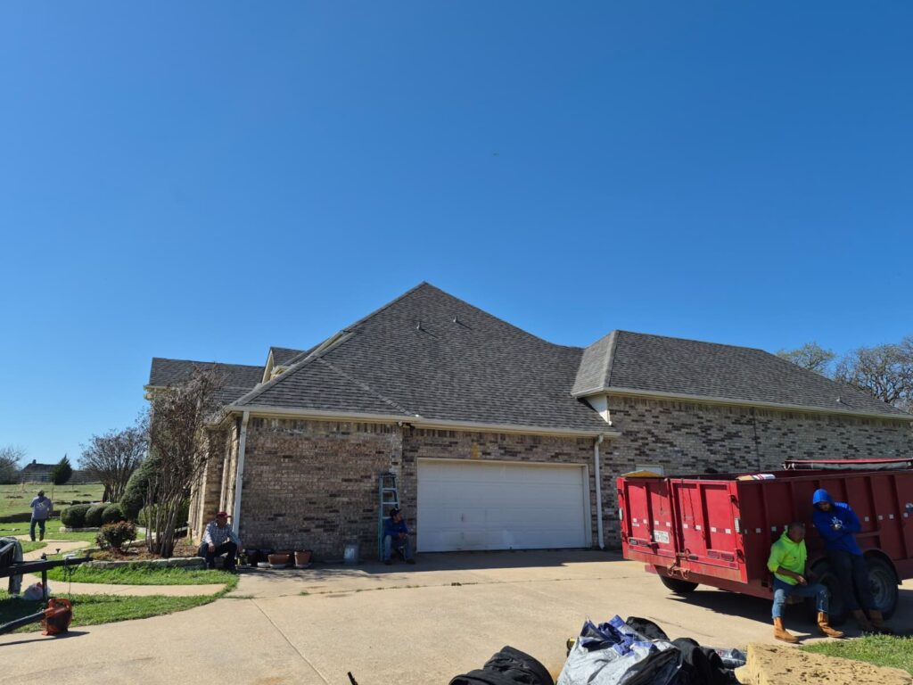 A residential house with a newly installed asphalt shingle roof is visible under a clear blue sky. Several workers are on the ground, some sitting by a red dump trailer and others near the garage.