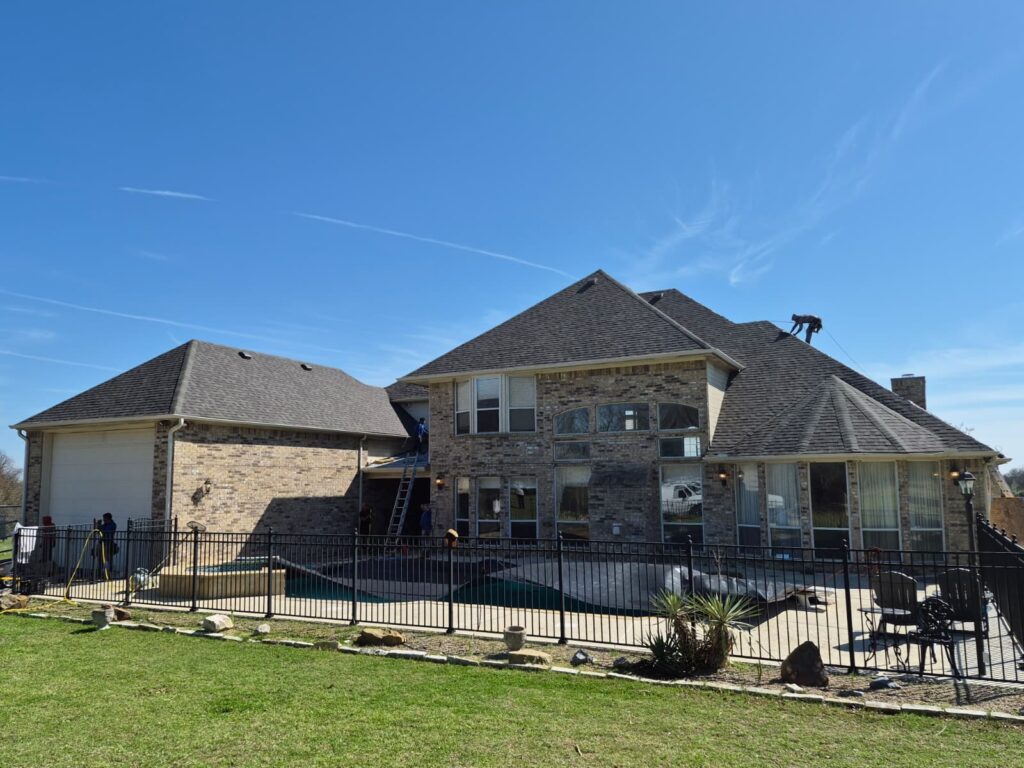 A large brick residential house with a multi-gabled asphalt shingle roof is undergoing work, with several crew members visible on the roof and around the property. A swimming pool and black metal fence are in the foreground under a clear blue sky.