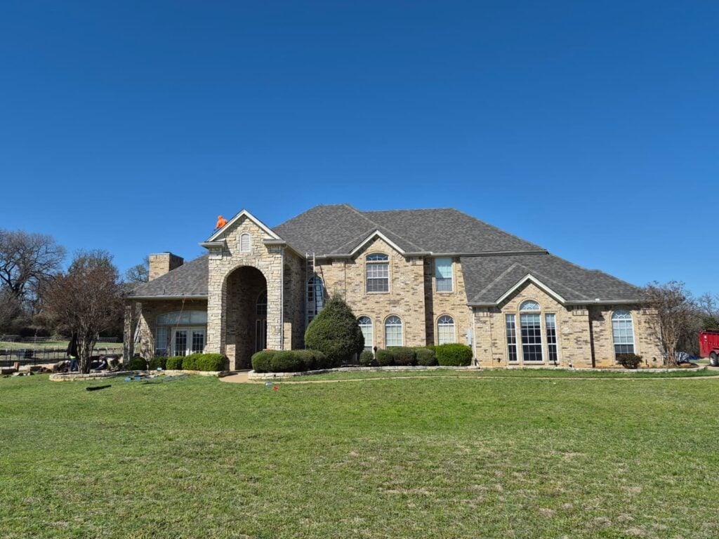 A large, two-story brick and stone house with a newly installed asphalt shingle roof under a clear blue sky. Several workers are visible on the ground to the left, and one worker in an orange shirt is on the roof near the front gable.