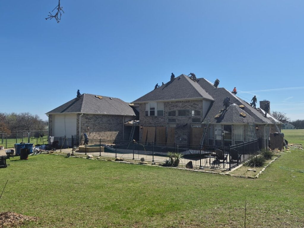 A large brick house with multiple roofers actively working on its dark gray asphalt shingle roof under a clear blue sky. The backyard features a swimming pool, patio furniture, and a grassy lawn.