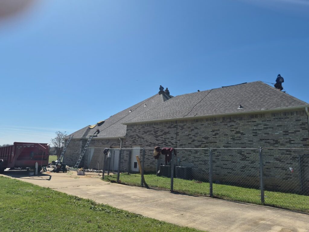 The image captures a residential brick house with a gray asphalt shingle roof, where several workers are actively engaged in roofing tasks. Ladders are propped against the house, and a red dumpster is visible in the driveway area.