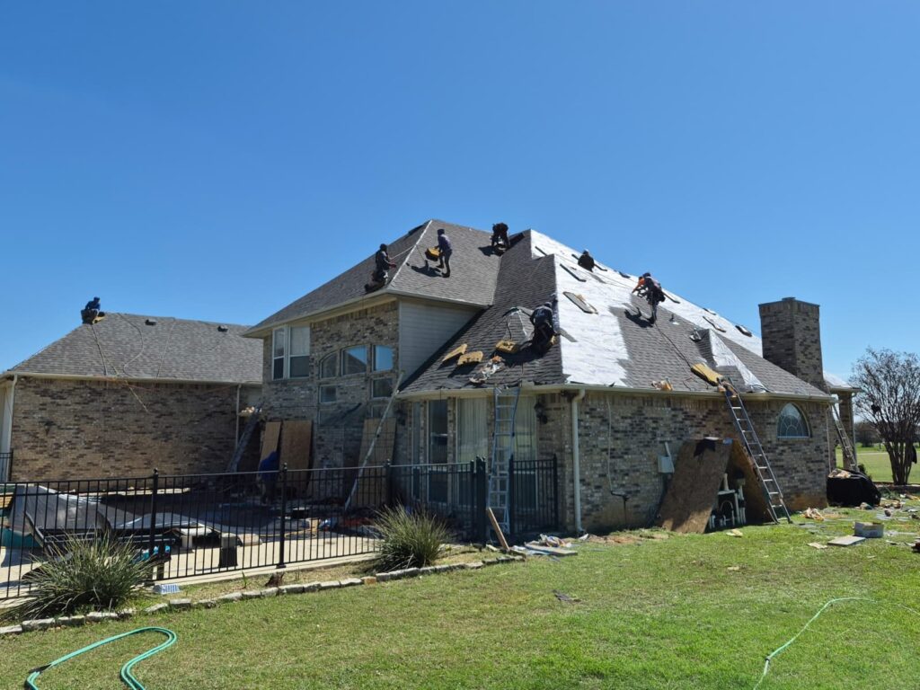 A crew of roofers is actively working on a large residential house, with sections of the old asphalt shingles removed and new underlayment visible, alongside areas where new shingles are being installed. Ladders and debris are visible around the perimeter of the house.