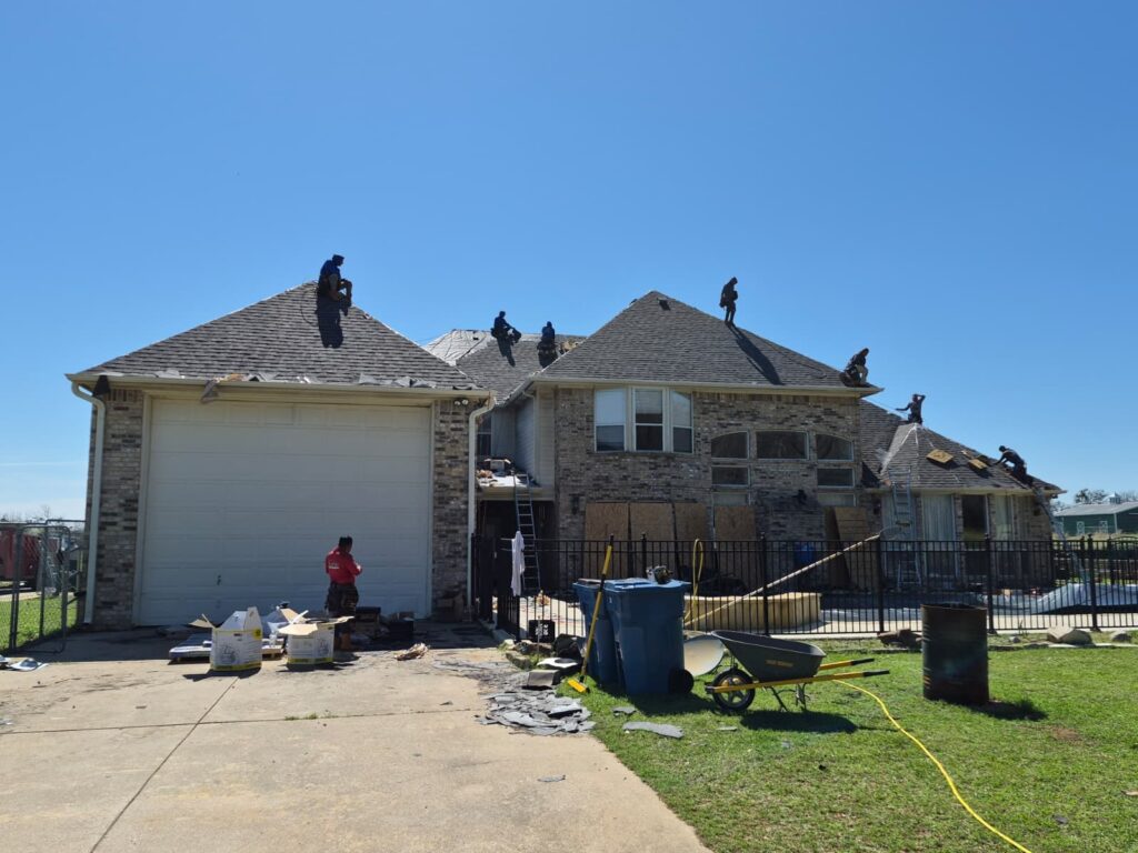 A large residential house with multiple roofers actively working on the asphalt shingle roof under a clear blue sky. Debris from the old roof is visible on the ground and driveway.
