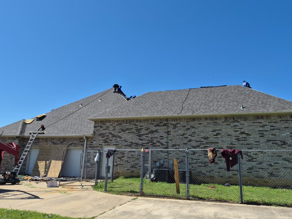 Several roofers are actively working on the asphalt shingle roof of a single-story brick residential house under a clear blue sky. A ladder is visible on the left side, and some roofing debris is on the ground.
