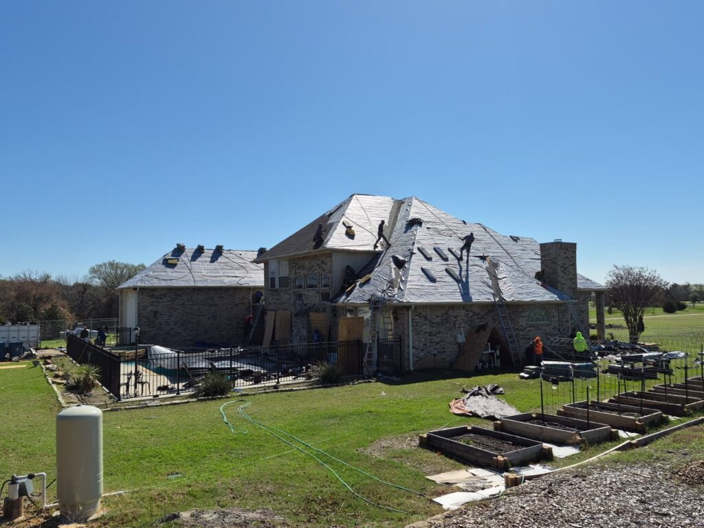 A large brick house with a complex roof structure is undergoing a roof replacement, with several crew members visible on the roof installing underlayment and new shingles. The surrounding yard includes a pool, garden beds, and various roofing materials and equipment.