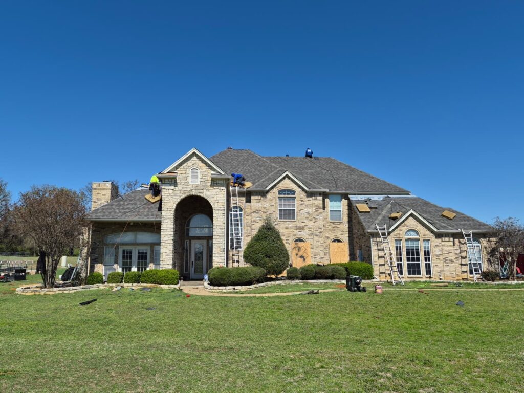 A large, two-story brick and stone house with a crew actively working on the asphalt shingle roof under a clear blue sky. Ladders are set up against the house, and some roofing materials are visible on the roof and ground.