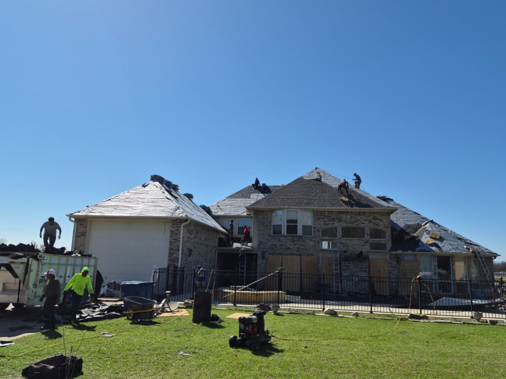 A large residential house undergoing a roof replacement, with multiple crew members actively working on the partially stripped and underlayment-covered roof sections. A dumpster is visible on the left, collecting old roofing materials.