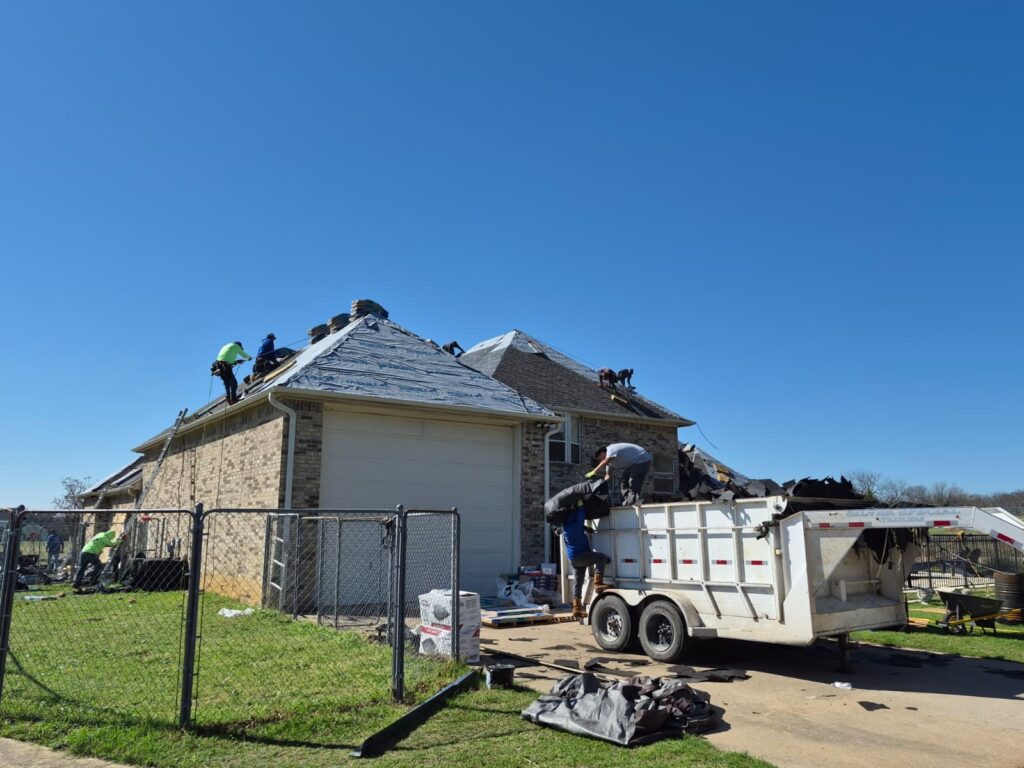 A crew of roofers is actively working on a residential brick house, with some sections of the roof stripped down to the underlayment and new shingle bundles visible. Workers are also seen loading old roofing debris into a large dump trailer parked in the driveway.