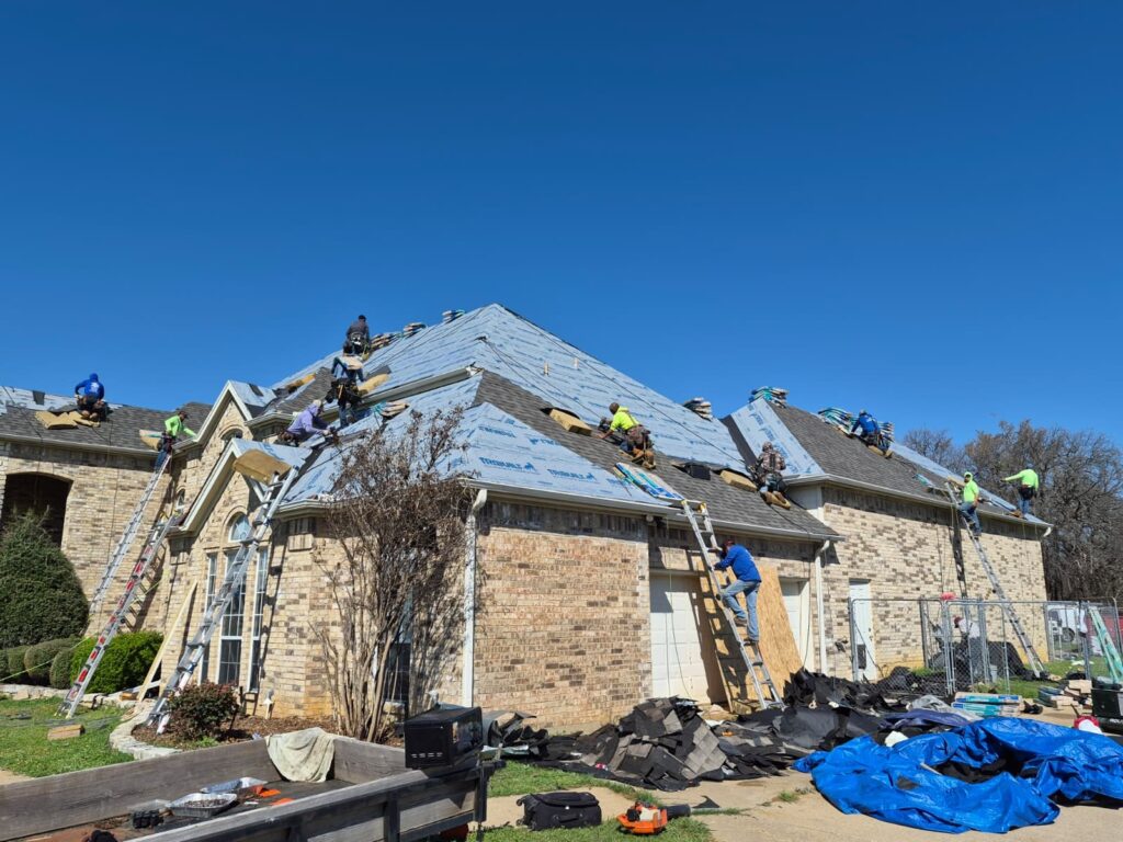 A crew of roofers is actively installing new asphalt shingles on a large brick residential house under a clear blue sky. Several workers are visible on different sections of the roof, with ladders and roofing materials present.