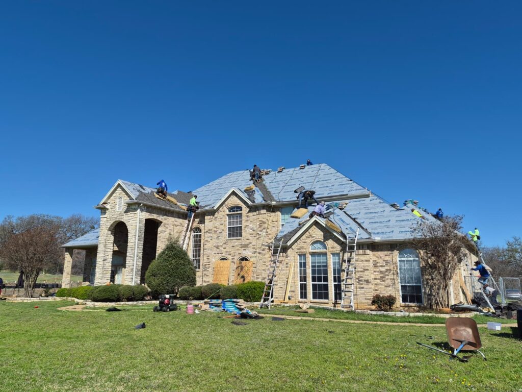 A large brick house with a complex roofline is shown with multiple roofers actively working, installing underlayment and new shingles. Ladders are positioned around the house, and roofing materials are visible on the ground.