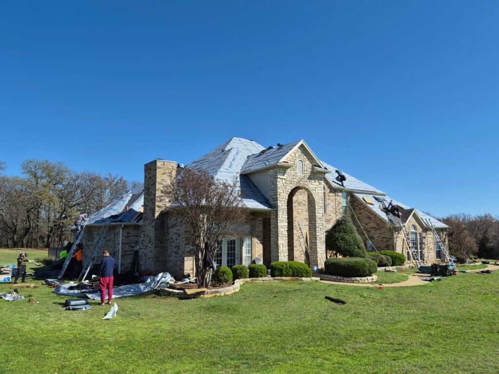 A large brick and stone house with a roofing crew actively working on replacing the roof. The old shingles have been removed, and new underlayment is visible, with some new asphalt shingles being installed.