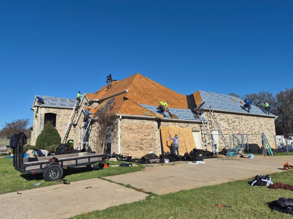 A crew of roofers is actively working on a large residential brick house under a clear blue sky. Parts of the roof are stripped to the decking, while other sections have new underlayment installed.