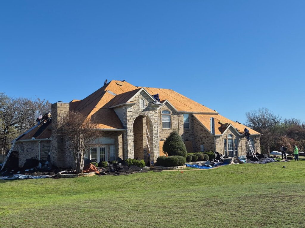 A large brick and stone residential house with a roofing crew actively working on the roof replacement. Old shingles are being removed, and new underlayment and some new shingles are visible, with debris collected on tarps around the perimeter.
