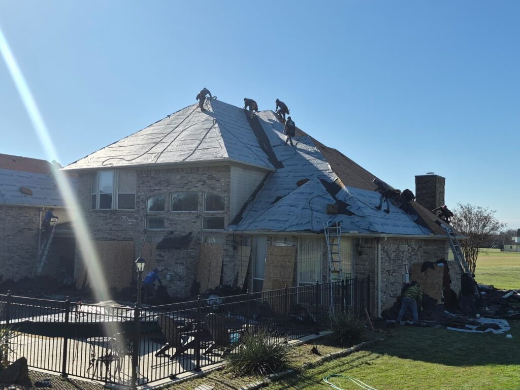 A crew of roofers is actively working on replacing the roof of a large brick house, with sections showing underlayment installed and new shingles being applied. Ladders are visible, and debris from the old roof is piled on the ground next to the house.