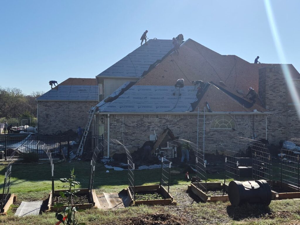 A crew of roofers is actively working on a large residential roof, installing underlayment and new asphalt shingles. Ladders are set up against the brick house, and ground protection is visible around the perimeter.