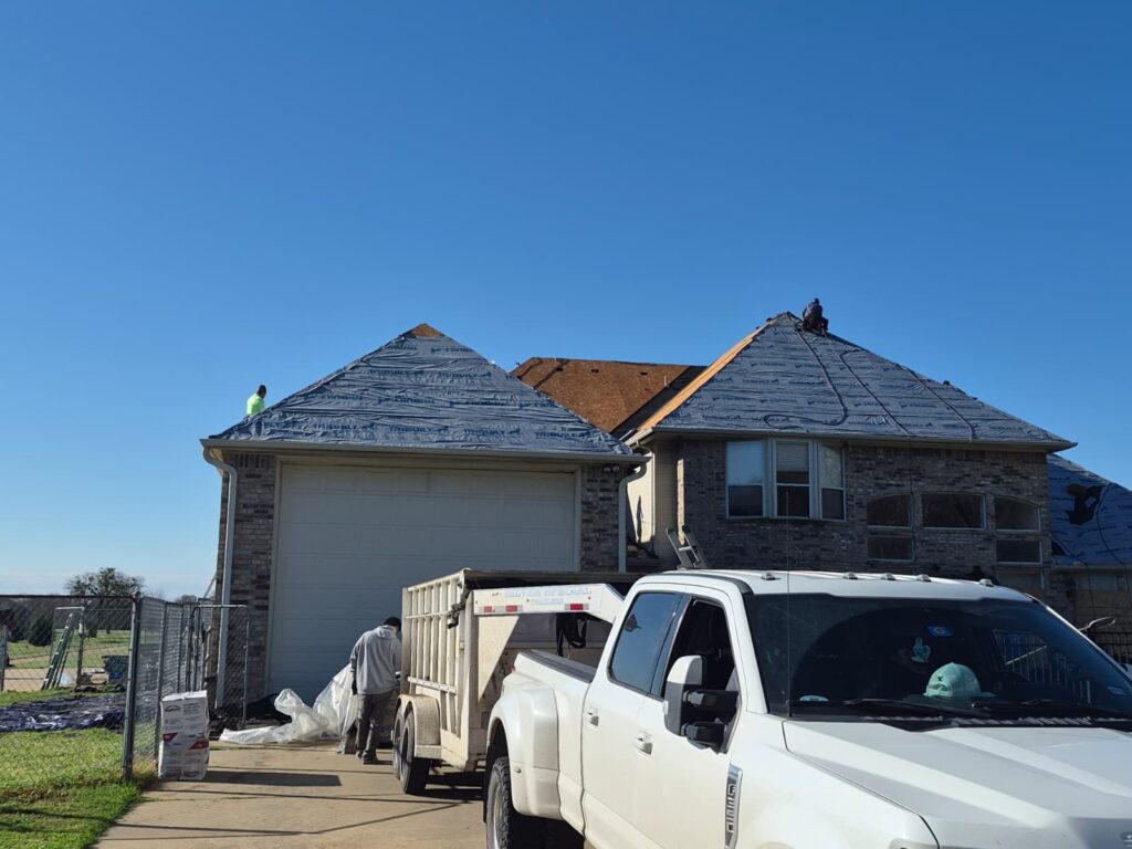 A residential house is shown during a roofing project, with blue underlayment covering most of the roof sections and some exposed decking. Several crew members are visible, one on the roof and another near a white pickup truck and trailer in the driveway.