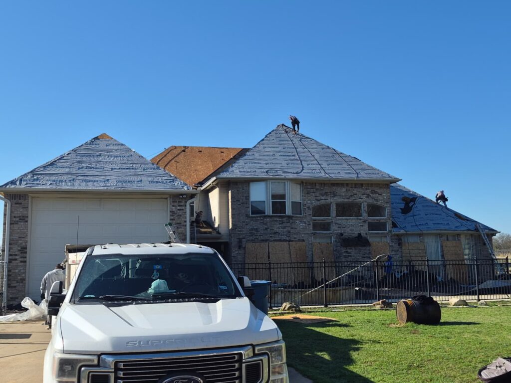 This image shows a large residential house undergoing roof replacement, with blue synthetic underlayment installed on most sections and workers actively on the roof. A white Ford Super Duty truck is parked in the driveway, partially obscuring the garage.