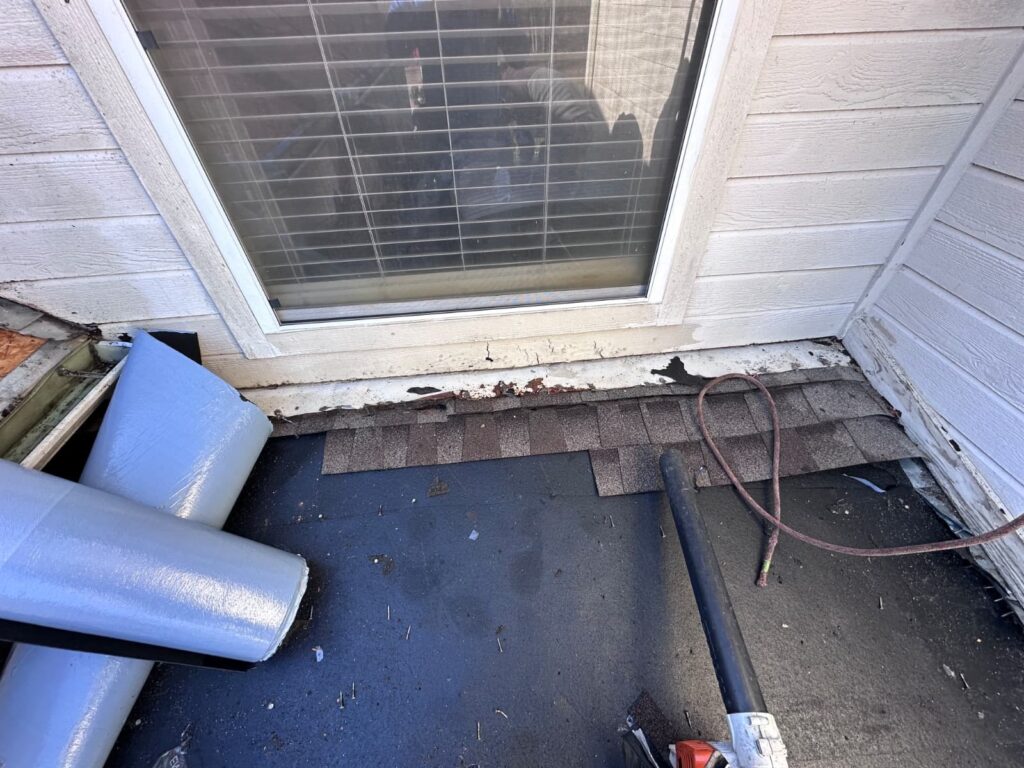 A close-up view of a roof section near a window, showing old asphalt shingles partially removed, exposing underlayment. Rolls of new underlayment or flashing material and a leaf blower are visible on the roof.