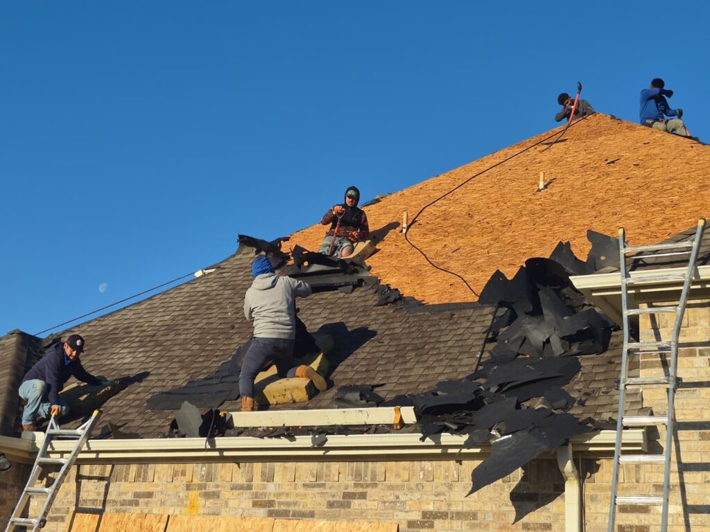Several roofers are actively removing old asphalt shingles from a residential roof, exposing the plywood decking underneath. A ladder is visible on the right, and the sky is clear blue with a faint moon.