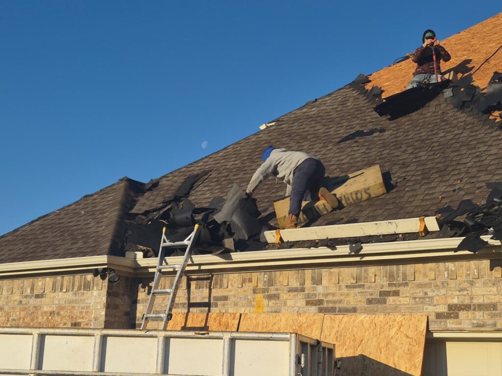 Two roofers are actively removing old asphalt shingles from a residential roof, with a ladder leaning against the house and a clear blue sky with a visible moon.